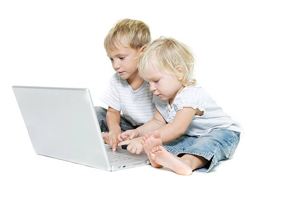 Two young children engaged in online learning with a laptop, surrounded by colorful notebooks and a cozy home setting.