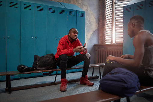 Two young African sportsmen sitting on bench and talking to each other after sports training in locker room