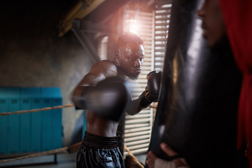 African muscular man in boxing gloves fighting with punching bag during sports training in gym