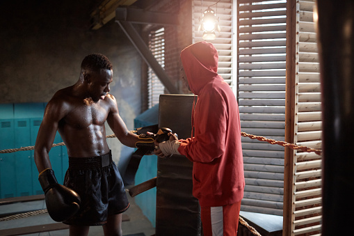 African muscular sportsman standing on boxing ring while his coach wearing boxing gloves on his hands