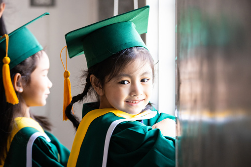 Asian elementary age girls wearing graduation caps and gowns smile while standing inside school.