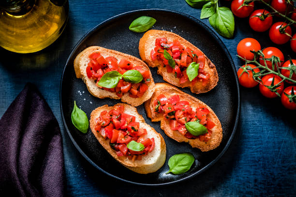 Italian bruschetta Top view of a homemade Italian bruschetta made with cherry tomatoes, basil, olive oil, garlic and salt disposed on a black plate surrounded by the ingredients for preparing the bruschetta on a dark blue backdrop. Low key DSLR photo taken with Canon EOS 6D Mark II and Canon EF 24-105 mm f/4L bruschetta stock pictures, royalty-free photos & images