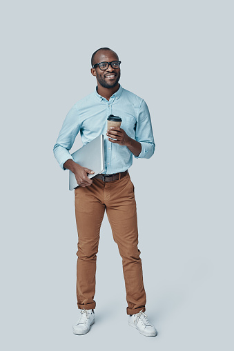 Full length of confident young African man carrying laptop and smiling while standing against grey background