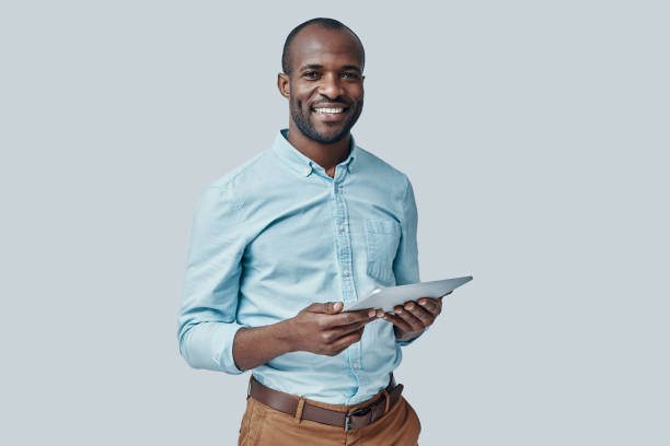 Happy young African man Happy young African man using digital tablet and smiling while standing against grey background macho photos stock pictures, royalty-free photos & images