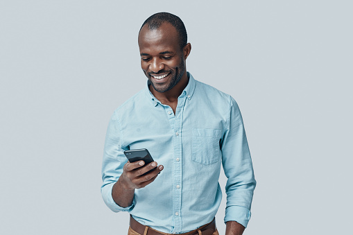 Handsome young African man using smart phone and smiling while standing against grey background