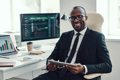 Concentrated young African man in formalwear using modern technologies and smiling while working in the office