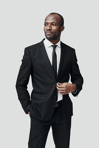 Stylish young African man in formalwear looking away and adjusting jacket while standing against grey background