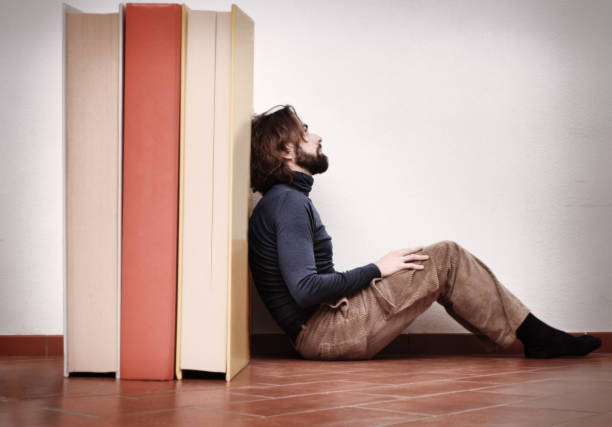 Small male figure in a suit reading a book while sitting on vibrant stacked books, creating a playful and creative scene.
