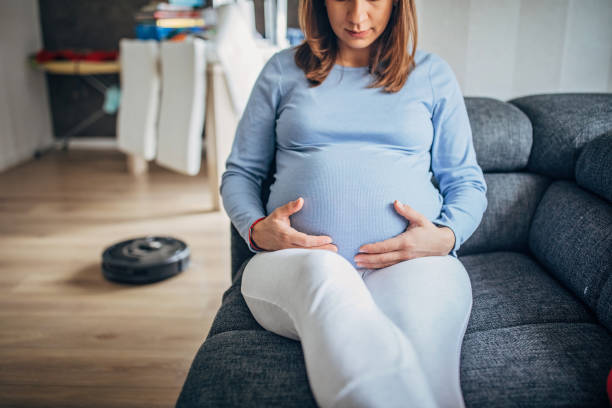 istock Pregnant lady sitting on sofa while robot vacuum cleaner is cleaning 1194673070
