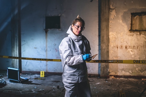 Female investigator in late 30s wearing protective clothing and looking at camera while making notes outside evidence area at crime scene.