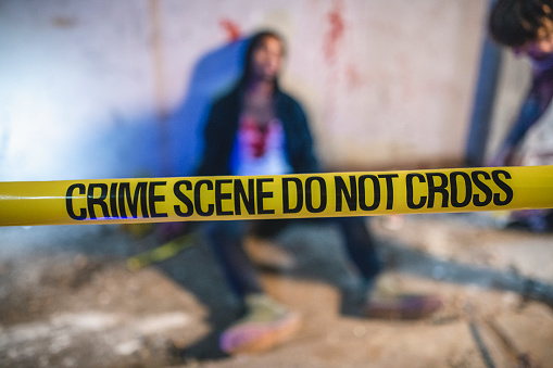 Focus on foreground crime scene cordon tape with blurred and bloody victim leaning against wall in background.