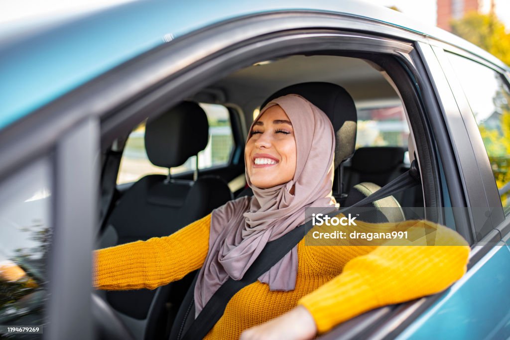 Beautiful woman with hijab driving a car. Young asian muslim woman in head scarf smile inside the car. Beautiful caucasian Muslim Woman wearing light hijab in a car lifestyle shoot. Beautiful muslim woman with toothy smile driving car. Car Stock Photo