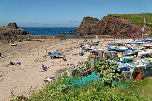 sandy beach with cliffs either side. Busy with holiday makers. Tents and windbreaks on the beach. Small yachts, fishing and rowing boats moored on the sand. Blue sea and sky.