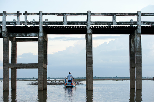 Boating, Ubein Bridge, Mandalay, Myanmar. A lethargic boatman rows his tourist boat through the arches of the teak bridge with tourists on board during a grey day on the lake, Ubein Bridge, Mandalay, Myanmar