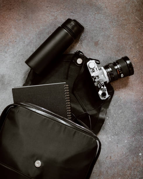 Black leather backpack or camera bag spilling out onto a concrete floor. A sustainable water bottle, vintage film camera, and spiral bound notebook are inside.
