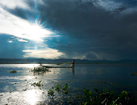 Inn Thar fisherman on Inle Lake, Shan State, Myanmar. A starburst of sunlight breaks through the cloud cover at dusk over Inle Lake as a 'one-legged' Inn Thar tribe fisherman spreads his traditional fishing nets onto the rippled reflective surface of the lake.