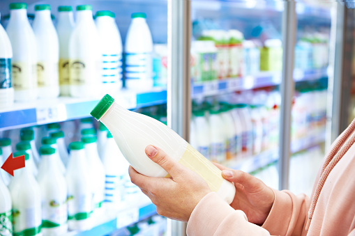 A smiling woman is reading the label on a bottle of fresh milk checking the sell-by date as she does her weekly shop in her local supermarket