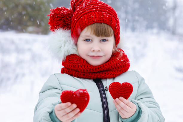 A slightly tenderly smiling small child in colorful clothes is showing two little knitted red hearts as symbol of love at the snowy forest background. The concept is St. Valentine's Day celebration.