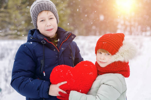 An brother with small sister dressed in warm clothes standing inside a snowy park are happily smiling and holding a heart together enjoing winter walking. The concept is St. Valentine day celebration.