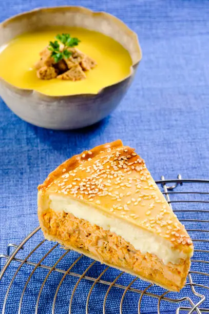 A bowl of vegetable soup with croutons on top on the background, and a slice of chicken pie with a crispy golden-brown crust and white sesame on a blue tablemat