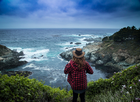 A young woman in a outdoorsy hat takes in a breathtaking view of the the pacific ocean and the incredible cliffs around it.