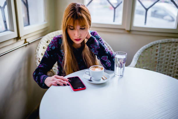 Young beautiful woman drinking coffee and looking at smartphone while sitting at cafeteria