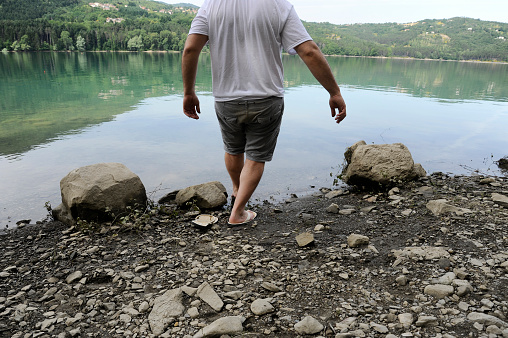 Man enters in the waters of mountain lake in the summer day. Man wearing jeans shorts, t-shirt and flip-flops. Calm surface of the water. Back view.