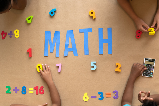 A group of multi-ethnic elementary students sit around a table covered in number blocks in a rainbow of colors.  The word 