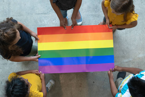 Aerial View of Multi-Ethnic Elementary Students Holding a Gay Pride Poster stock photo A group of multi-ethnic elementary students stand around a colorful gay pride poster.  They are each holding an edge of the poster so it is out flat for the camera to see.  Only the tops of their heads can be seen as the picture is taken from an aerial view.  Each of the students are wearing casual clothing. lgbtqia rights photos stock pictures, royalty-free photos & images