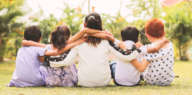 Back view, Group of kindergarten multi ethnic kids friends arm around sitting together at park outdoor - Concept showing of childhood friendship, togetherness with diversities. Back view, Group of kindergarten multi ethnic kids friends arm around sitting together at park outdoor - Concept showing of childhood friendship, togetherness with diversities arm around stock pictures, royalty-free photos & images