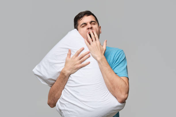 istock Tired brunet man in a blue tee yawning hugging white pillow isolated over grey background. 1194812615