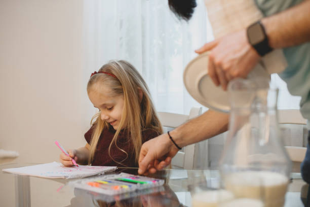 прекрасная маленькая девочка рисует на кухне, в то время как ее отец моет посуду. - family with one child domestic life caucasian love стоковые фото и изображения