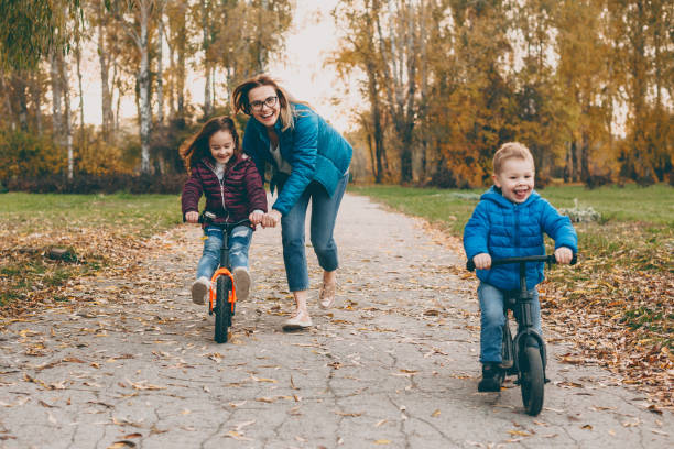 charming young mother playing with her daughter and son riding bicycle while boy is going . mother helping her little girl to ride a bicycle. - smiling little girls little boys autumn imagens e fotografias de stock