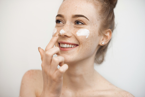 Retrato de una joven encantadora con el pelo rojo y pecas aisladas sobre blanco aplicando una crema anti edad en la cara y la nariz sonriendo. photo