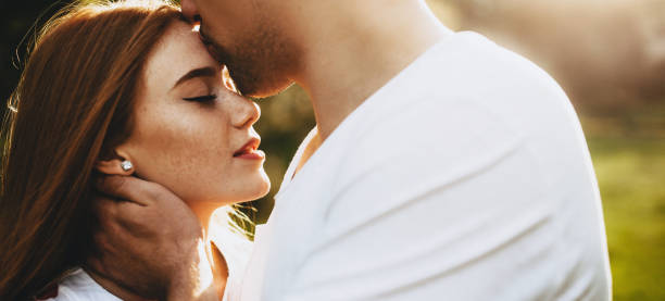 Side view of a charming young couple while man is kissing on forehead her red haired girlfriend which is with closed eyes against sunset.
