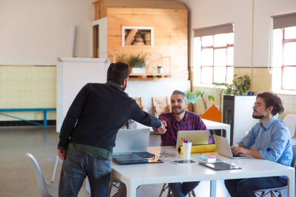 istock Startup team sitting together, using laptops, greeting coworker 1194828072