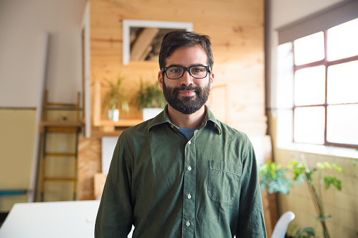 Positive hipster entrepreneur, IT expert, software developer posing in modern office. Bearded young man in glasses standing in contemporary office space. Corporate portrait concept