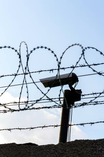 istock Security camera behind barbed wire fence on the wall, prison, security, crime or illegal immigration concept, blue sky background 1194836489