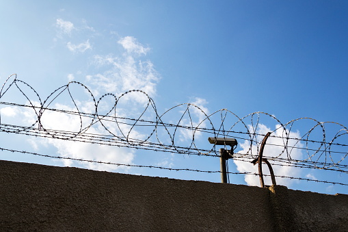 Security camera behind barbed wire fence on the wall, prison, security, crime or illegal immigration concept, blue sky background