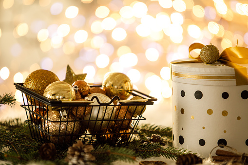 Shot of basket full of golden ornaments set on a tree branch next to a beautifully decorated Christmas gift box with a background of fairy lights.