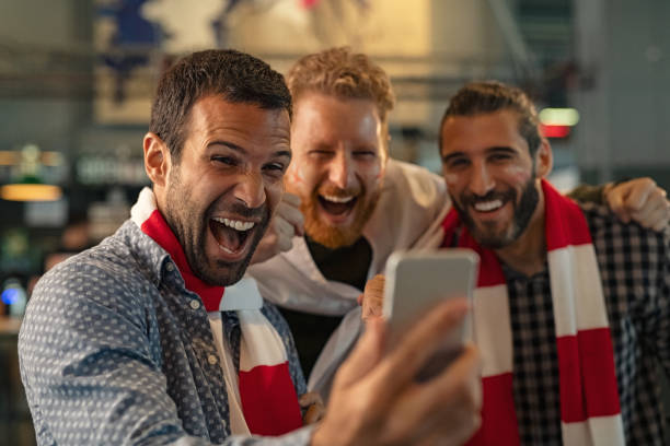Excited supporters watching football match on phone Excited men watching football in streaming on smartphone in bar. Football fans watching game on smart phone and celebrating victory score at pub. Happy supporters cheering and exulting after winning an online bet. fan enthusiast stock pictures, royalty-free photos & images