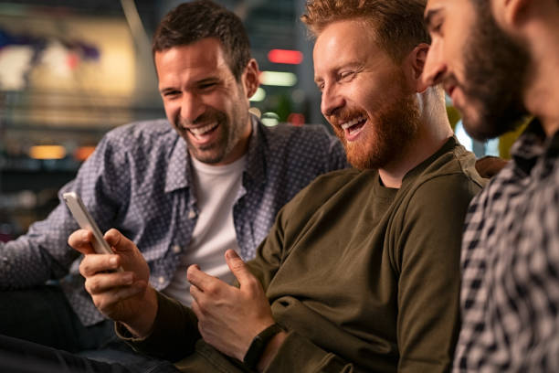 Friends laughing and using smartphone at pub Friends meeting in bar, using smartphone and spending time together. Happy young friends sitting on couch and watching streaming on mobile phone while laughing. Mid adult man with friends holding smart phone while watching funny video. friends in bar with phones stock pictures, royalty-free photos & images