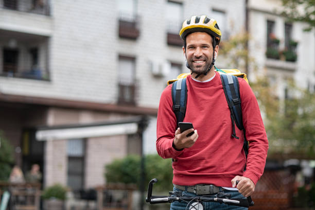 Food delivery boy on bicycle Delivery food courier guy checking order via mobile phone. Smiling man with delivery bag holding smartphone and wearing helmet on cycle. Food delivery courier with yellow backpack riding bicycle on city street with copy space. Fast lunch takeaway delivery. cycling helmet stock pictures, royalty-free photos & images