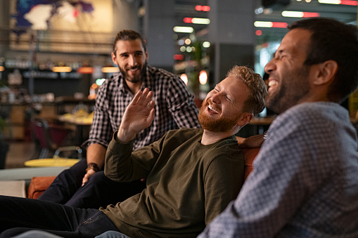 Group of three best friends laughing and enjoying the evening at pub. Happy young men enjoying late night staying together at bar. Cheerful guys sitting on couch and having fun while relaxing after work.