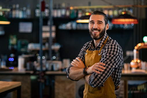 Happy satisfied bartender wearing apron while standing near counter with crossed arms and looking at camera. Confident coffee shop waiter smiling at cafeteria. Successful proud young brewer at his pub with copy space.