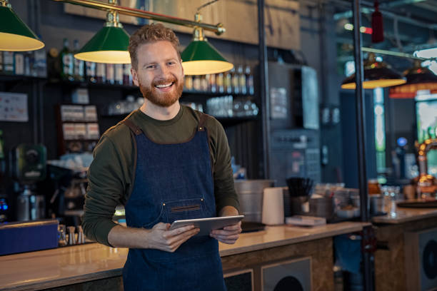 Smiling Waiter ready to take order at pub Successful small business owner using digital tablet and looking at camera. Happy smiling waiter wearing apron and holding digital tablet ready to take order. Portrait of young entrepreneur of coffee shop standing at counter with copy space. distillery photos stock pictures, royalty-free photos & images
