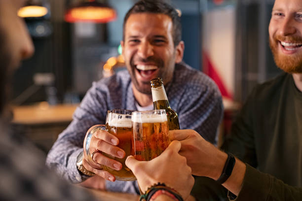 Friends toasting beer glass and bottle Happy mid adult friends clinking with beer mugs in pub. Three cheerful guys drinking draft beer, celebrating meeting and smiling. Laughing young men enjoying cold pint of beer during night at bar. beer stock pictures, royalty-free photos & images