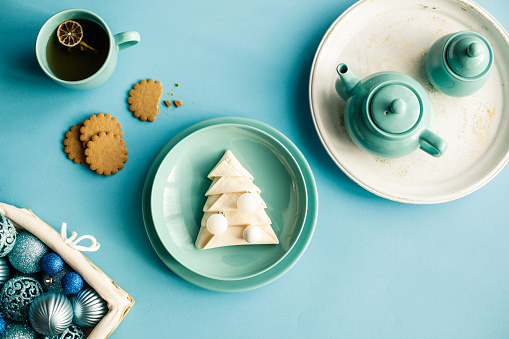 Directly above shot of a turquoise colored background with a plate in the middle with a delicious cookie shaped like a Christmas tree, surrounded with basket full of ornaments, gingerbread cookies and tea.