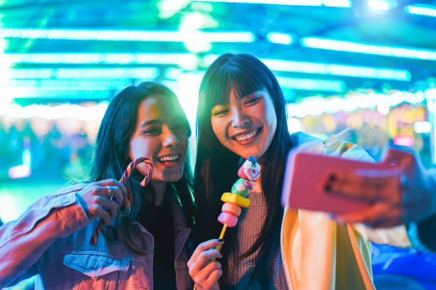 filles asiatiques heureuses mangeant des bonbons et prenant le selfie au parc d'attractions - jeunes amis à la mode ayant l'amusement avec la tendance de technologie - technologie, amitié et concept d'influenceur - foyer sur le visage féminin droit - a la mode photos et images de collection