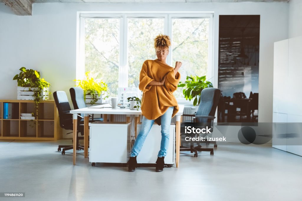 Confident woman standing at startup office Full length portrait of confident female business professional standing at her desk. Woman in casuals standing in office and looking at camera. Hipster Culture Stock Photo